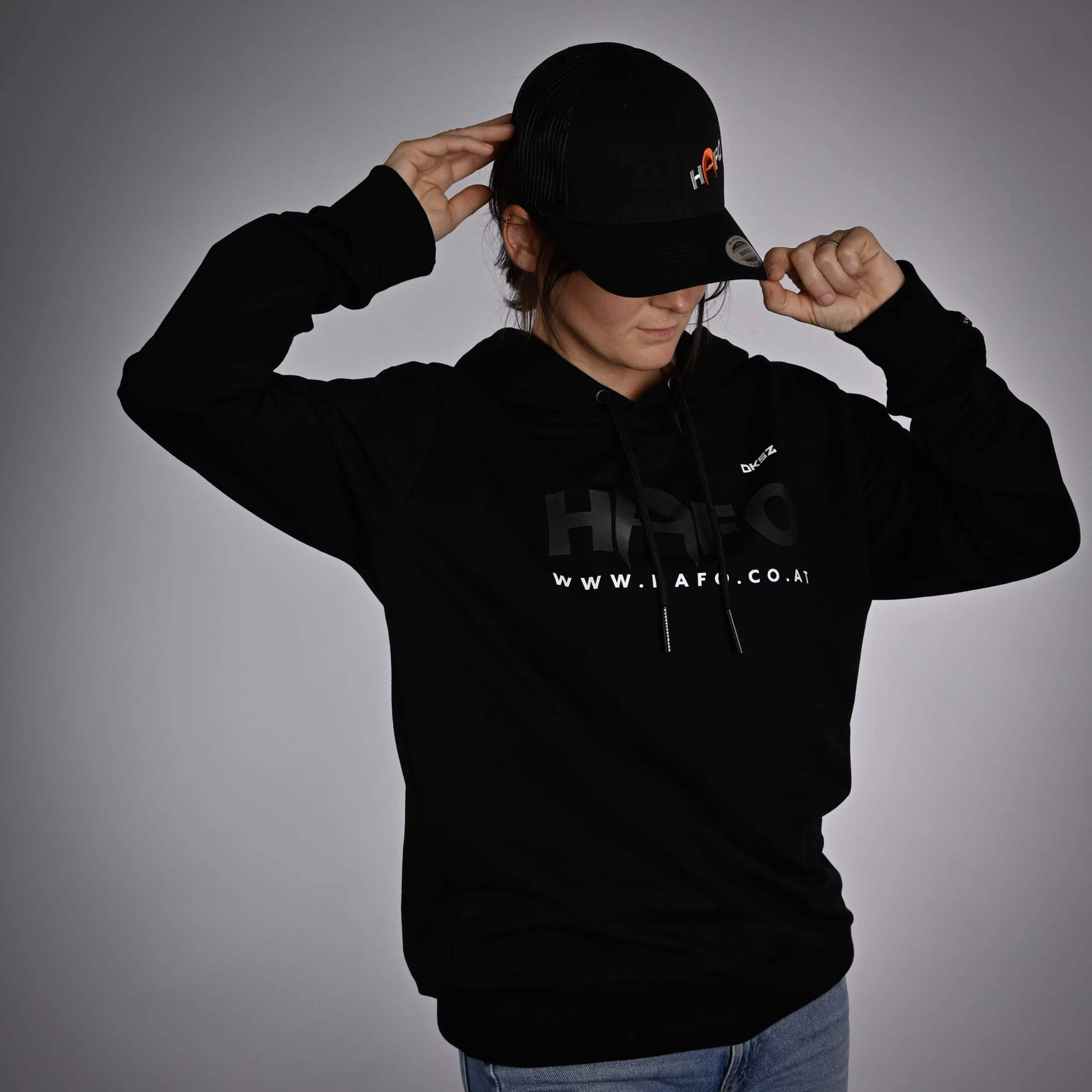 Eine Frau steht frontal zur Kamera im schwarzen HAFO DUNKELSCHWARZ Hoodie. Vorne sind die Aufdrucke „HAFO“, „DKSZ“ und ein Website-Link zu sehen. Sie trägt eine HAFO Kappe, die sie mit den Händen hält.