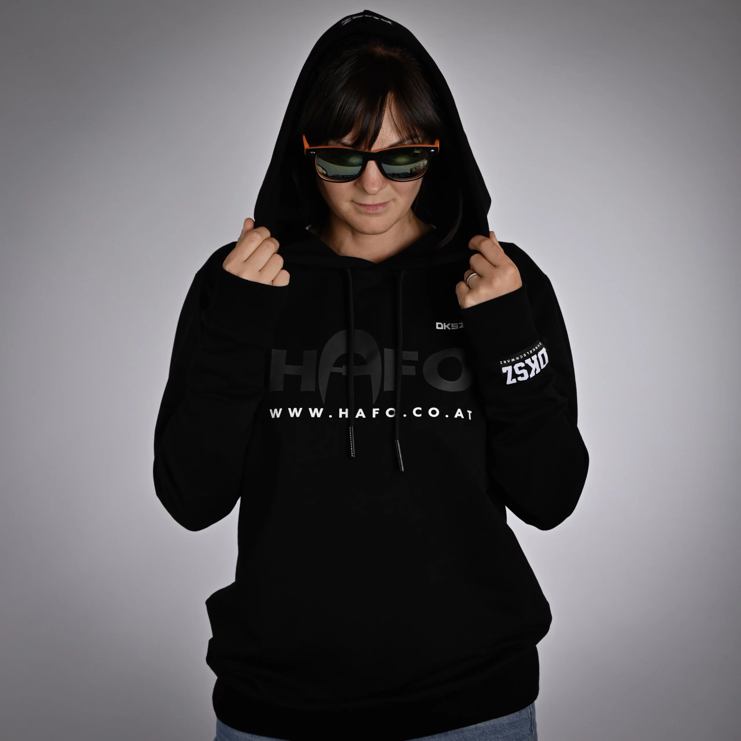Eine Frau steht frontal zur Kamera, trägt den schwarzen HAFO DUNKELSCHWARZ Hoodie mit aufgesetzter Kapuze. Sie hält die Kapuze mit den Händen, trägt eine Sonnenbrille und schaut auf den Boden. Auf dem Hoodie sind HAFO, eine Webadresse und DKSZ vorne sowie DKSZ am Ärmel sichtbar.